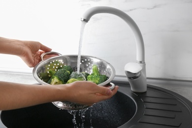 Photo of Woman washing fresh green broccoli in metal colander under tap water, closeup