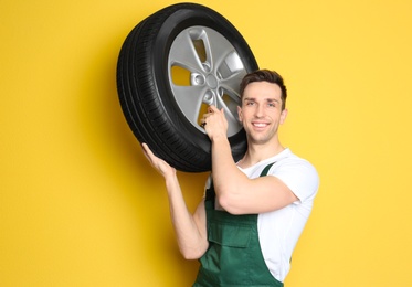 Photo of Young mechanic in uniform holding car tire on color background