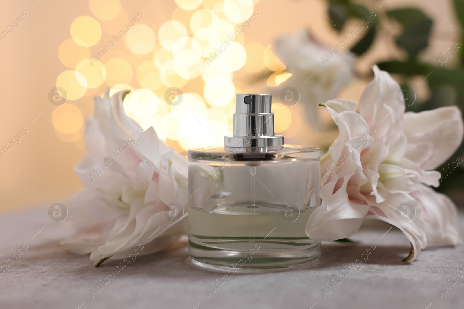 Photo of Bottle of perfume and beautiful lily flowers on table against beige background with blurred lights, closeup