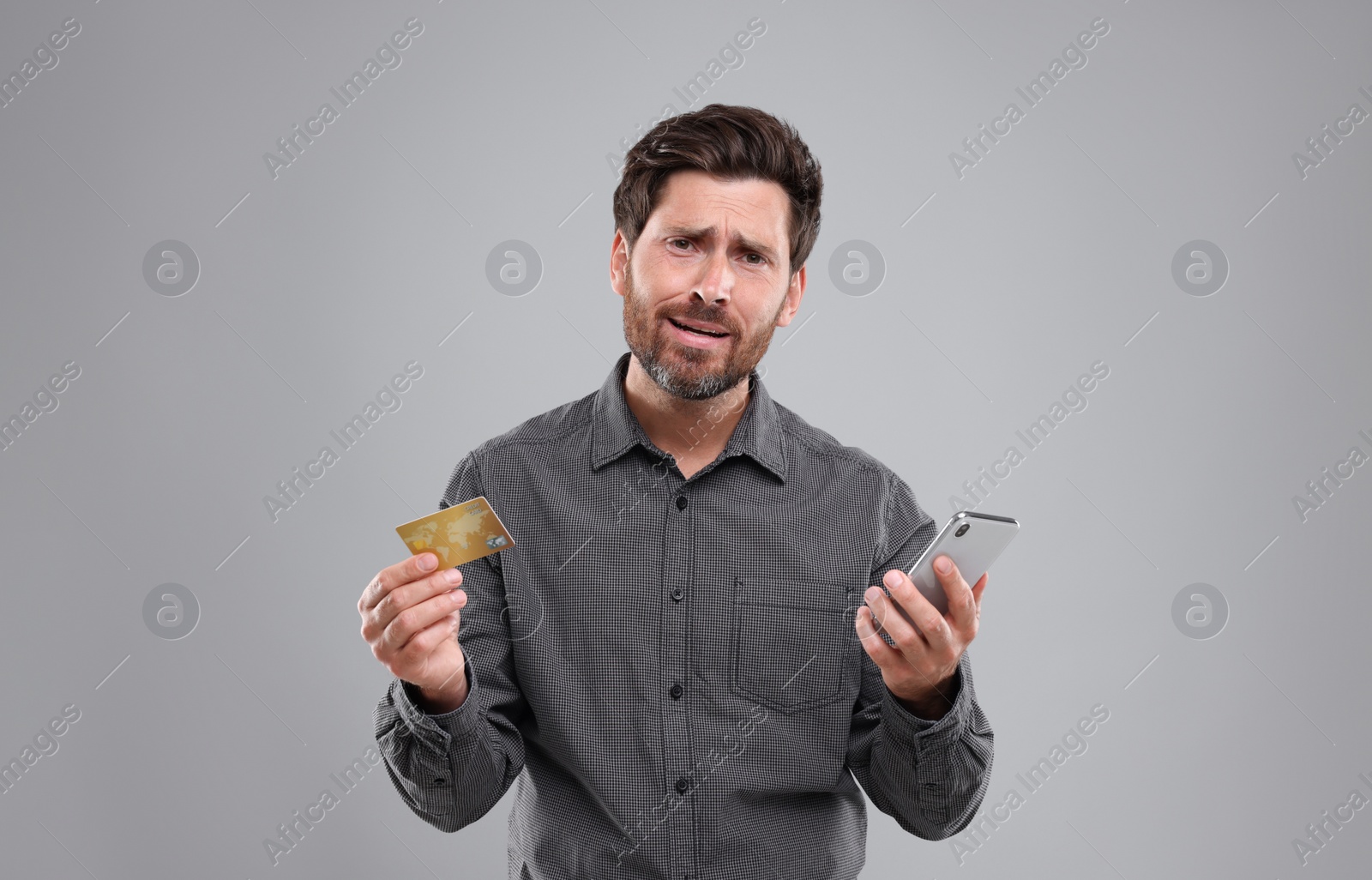 Photo of Upset man with smartphone and credit card on light grey background. Be careful - fraud