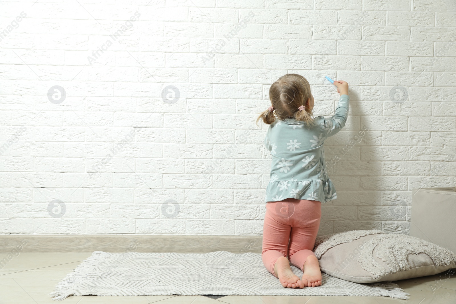 Photo of Little girl drawing on white brick wall indoors, back view and space for text. Child`s art