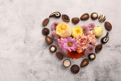 Heart made with delicious chocolate candies and beautiful flowers on grey table, flat lay. Space for text