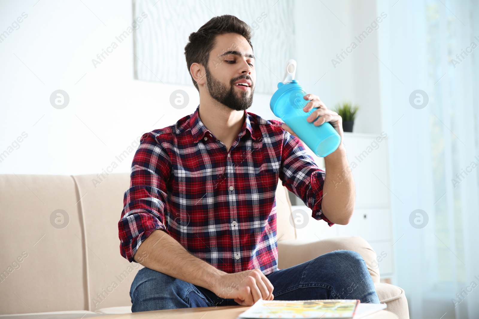 Photo of Young man with bottle of protein shake sitting on sofa at home