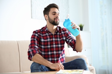 Young man with bottle of protein shake sitting on sofa at home