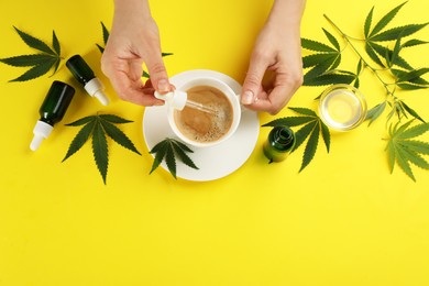 Top view of woman dripping THC tincture or CBD oil into coffee on yellow background, closeup. Space for text