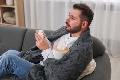 Photo of Sick man wrapped in blanket with tissue on sofa at home. Cold symptoms