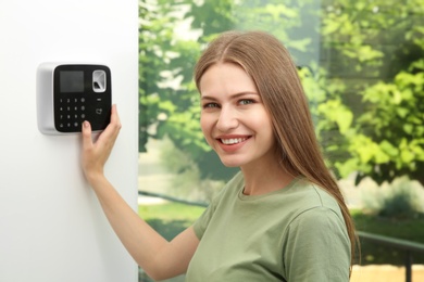 Photo of Young woman entering code on alarm system keypad indoors