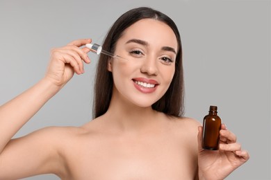 Happy young woman applying essential oil onto face on light grey background