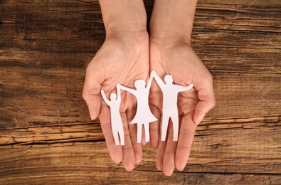 Photo of Woman holding paper figures of happy family in hands on wooden background, top view
