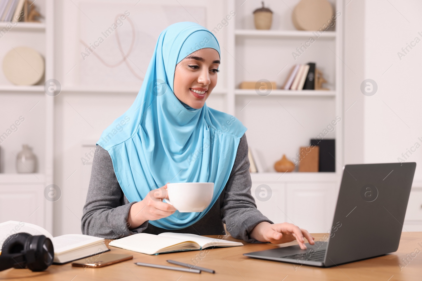 Photo of Muslim woman in hijab with cup of coffee using laptop at wooden table in room