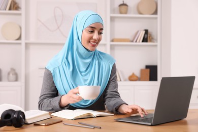Muslim woman in hijab with cup of coffee using laptop at wooden table in room