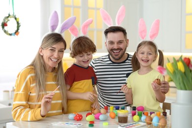 Photo of Painting Easter eggs. Happy family with bunny ears at white marble table in kitchen