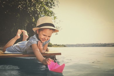 Image of Cute little girl playing with paper boat on wooden pier near river. Retro photo effect 