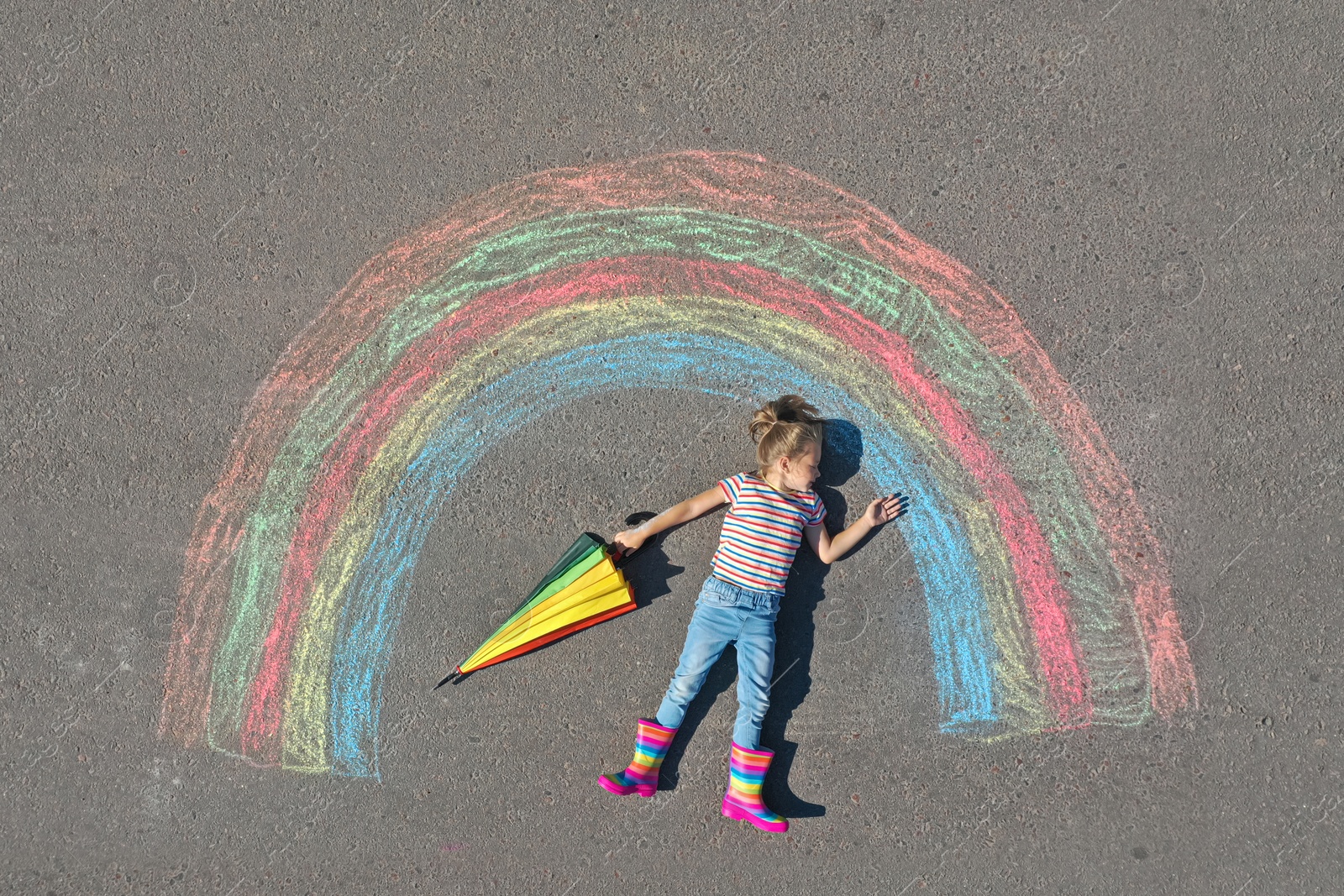 Photo of Cute little child with umbrella lying near chalk drawing of rainbow on asphalt, top view