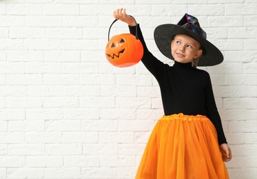 Photo of Cute little girl with pumpkin candy bucket wearing Halloween costume near white brick wall. Space for text