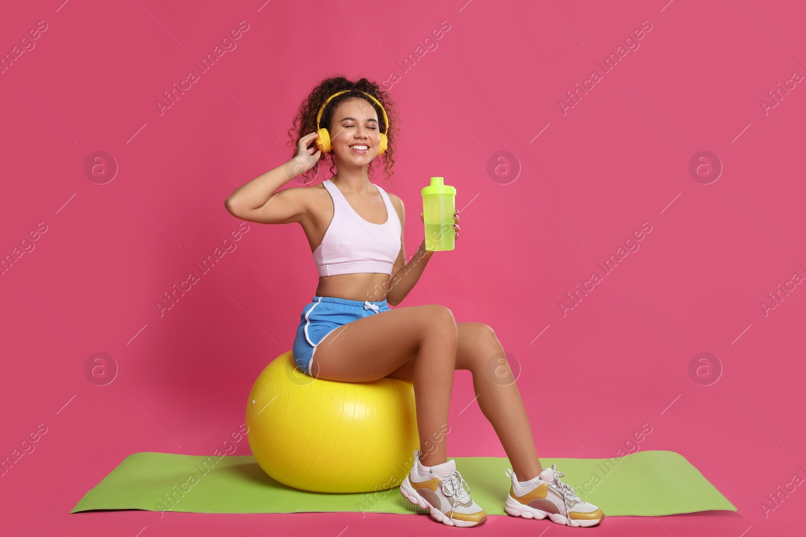Photo of Beautiful African American woman with headphones  on yoga mat against pink background