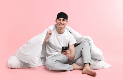 Happy man in pyjama with sleep mask, blanket and smartphone on pink background