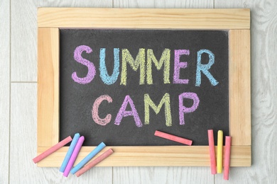 Blackboard with text SUMMER CAMP chalked in different colors on wooden background, top view