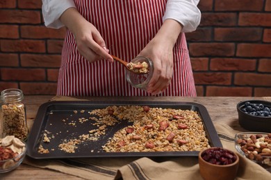 Photo of Woman putting granola from baking tray into jar at wooden table, closeup