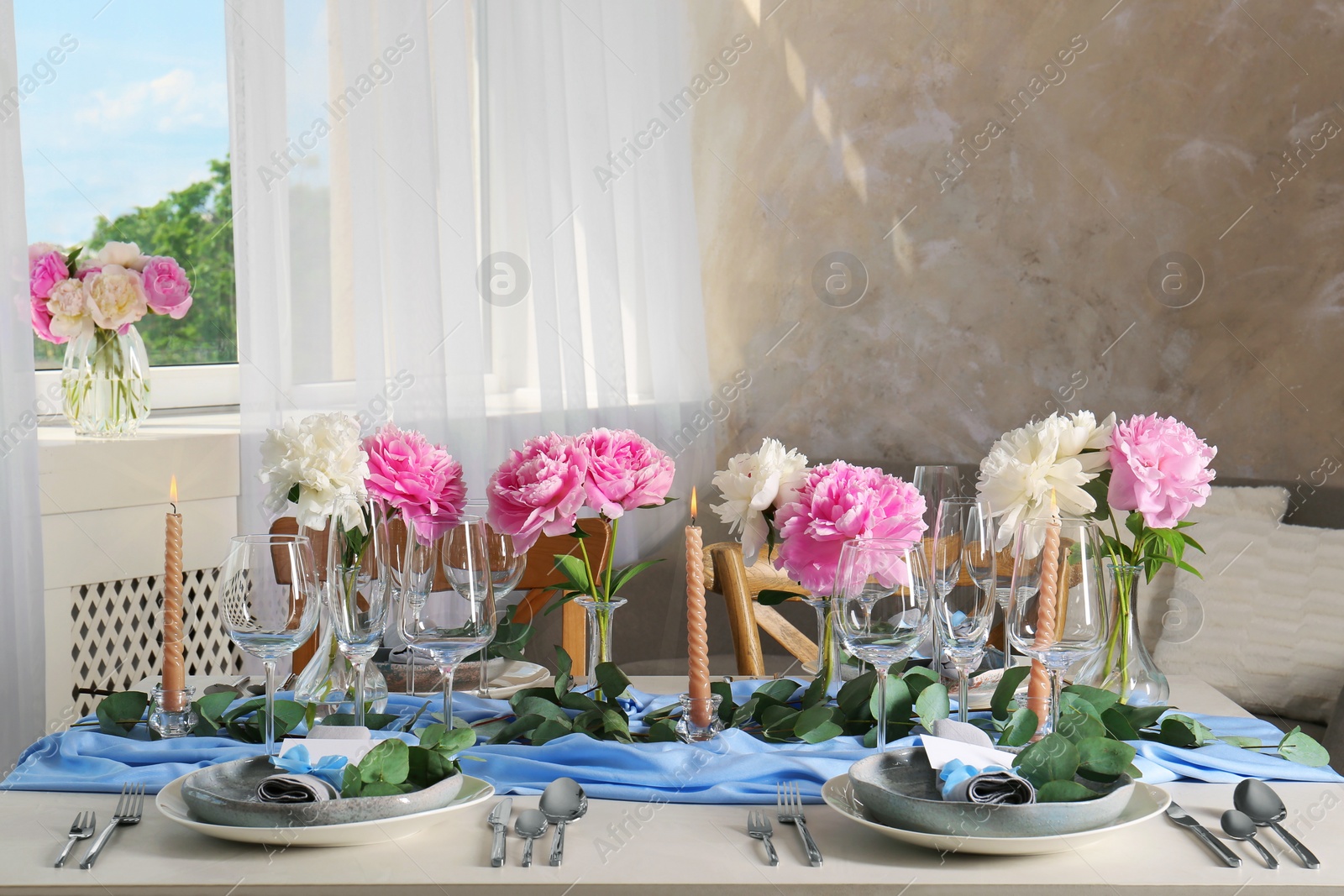 Photo of Beautiful table setting. Plates with greeting cards, napkins and branches near glasses, peonies, burning candles and cutlery on table in room