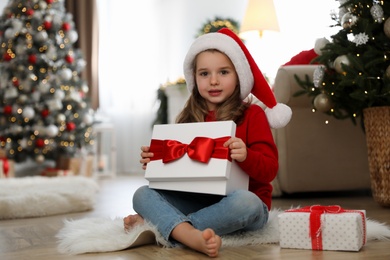 Photo of Cute little girl opening gift box in room decorated for Christmas