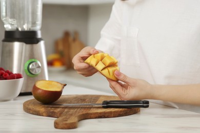 Woman preparing mango for tasty smoothie at white marble table in kitchen, closeup