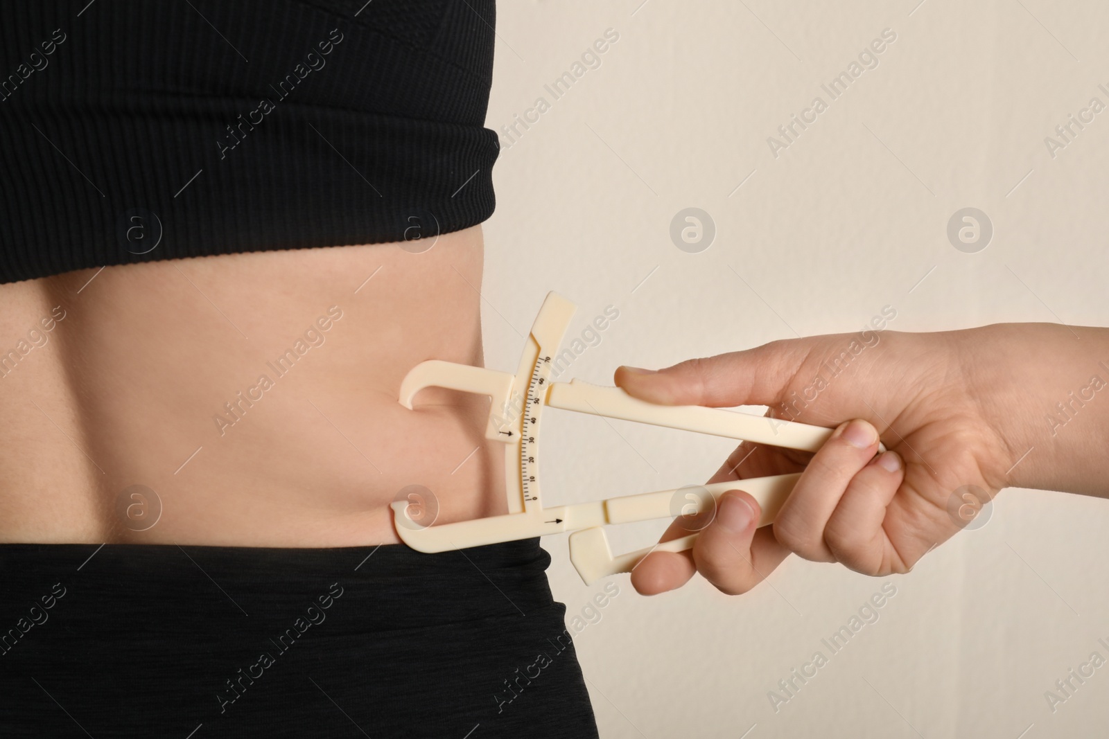 Photo of Nutritionist measuring woman's body fat layer with caliper on beige background, closeup