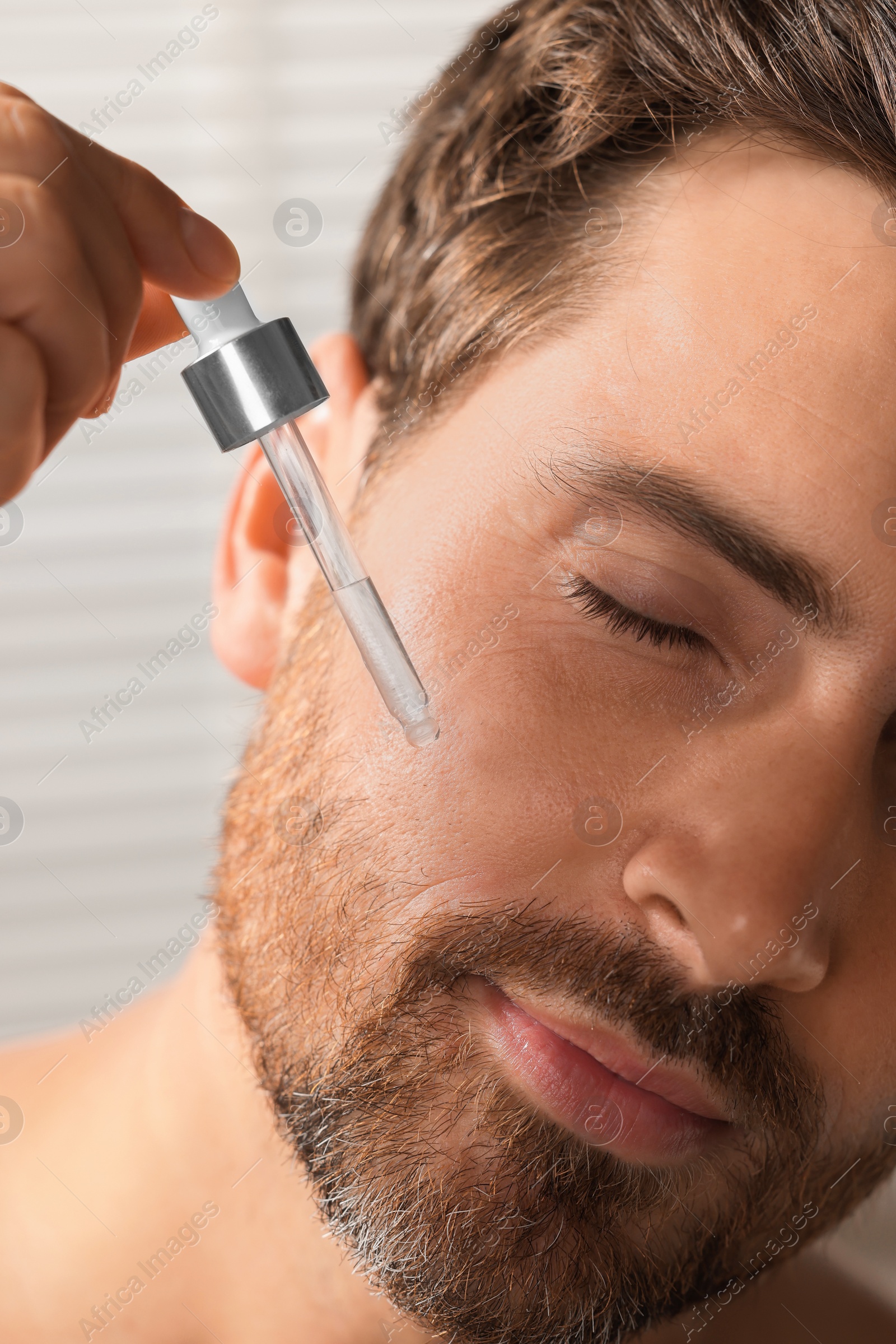 Photo of Handsome man applying cosmetic serum onto his face indoors, closeup