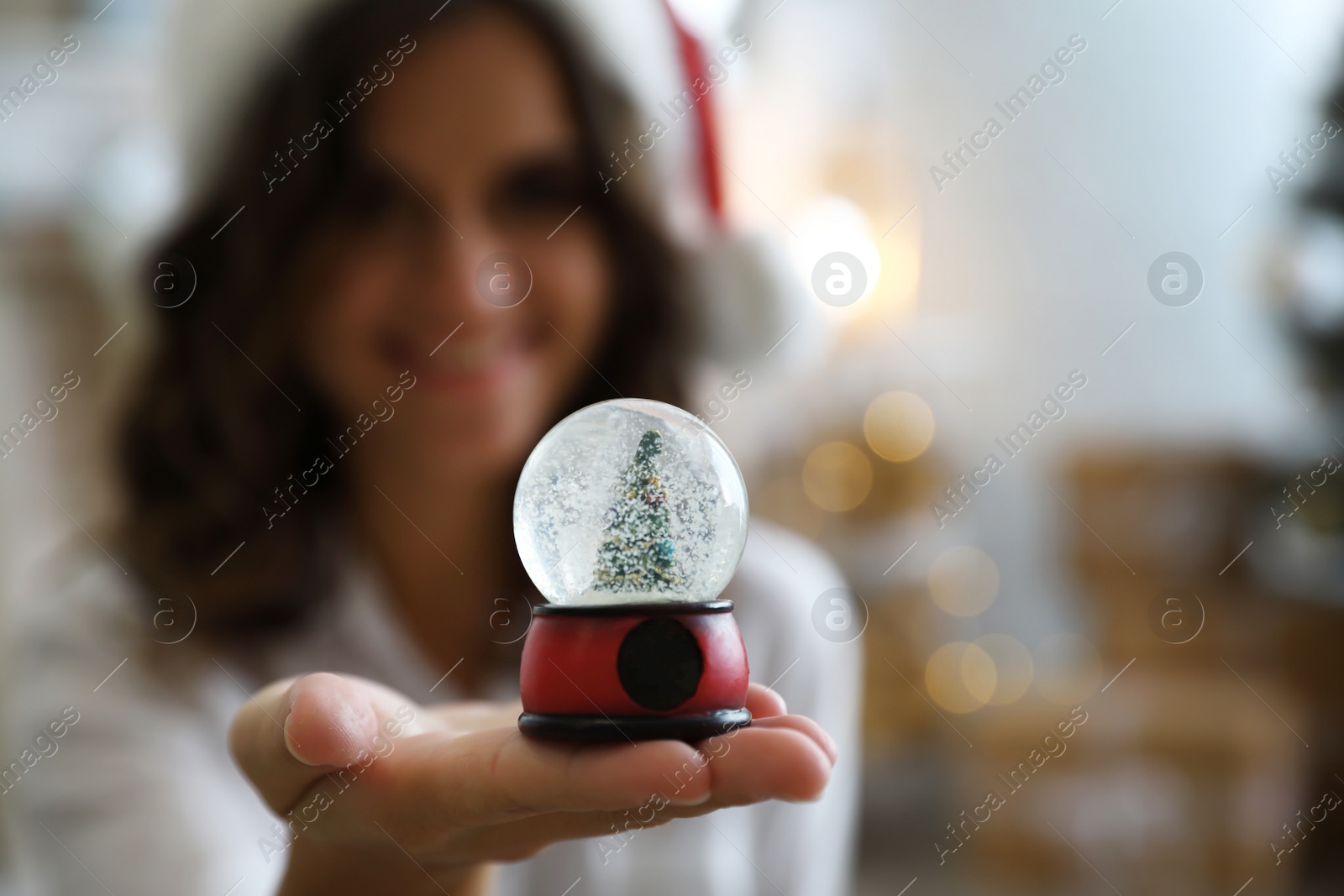 Photo of Young woman wearing Santa hat indoors, focus on hand with Christmas snow globe