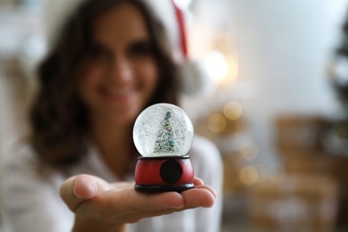 Young woman wearing Santa hat indoors, focus on hand with Christmas snow globe