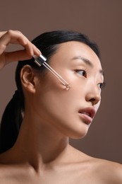 Beautiful young woman applying cosmetic serum onto her face on brown background