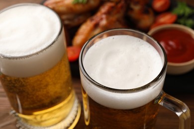 Mugs with beer, delicious baked chicken wings and sauce on table, closeup