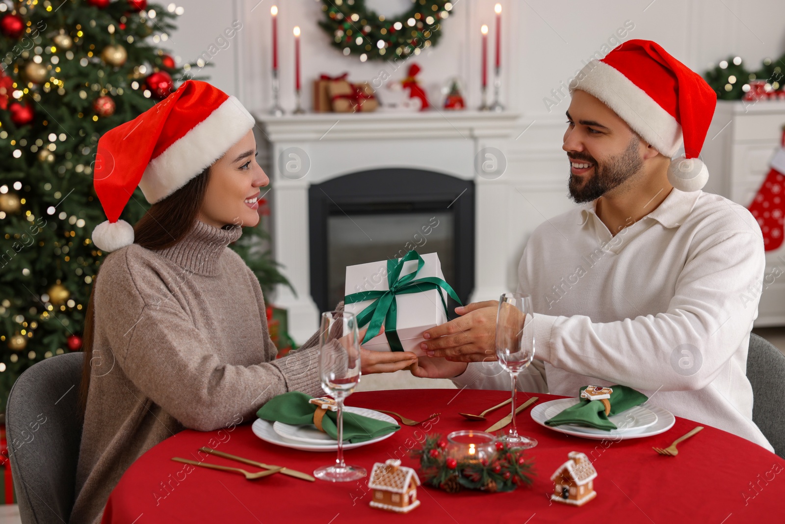 Photo of Beautiful young woman in Santa hat presenting gift to her boyfriend at table served for Christmas