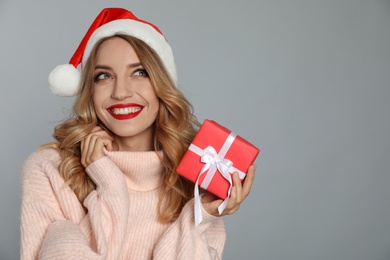 Photo of Happy young woman wearing Santa hat with Christmas gift on grey background. Space for text