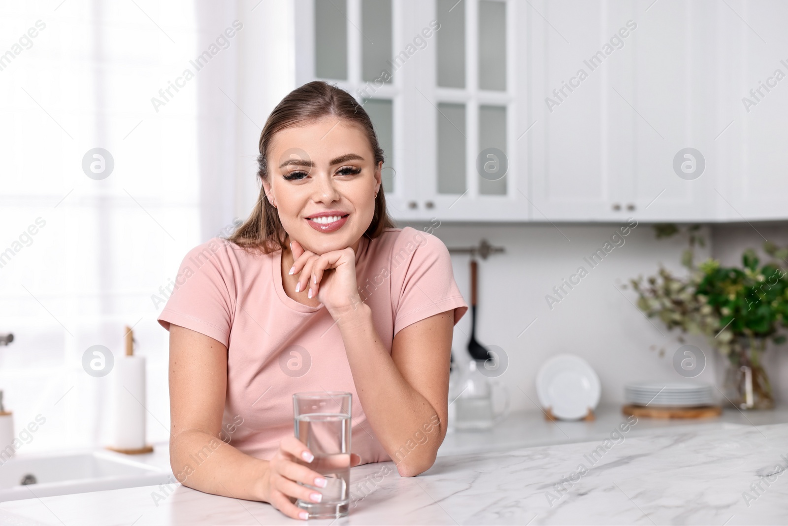 Photo of Happy woman with glass of fresh water at white marble table in kitchen, space for text