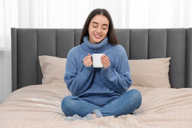 Photo of Happy young woman holding white ceramic mug on bed at home