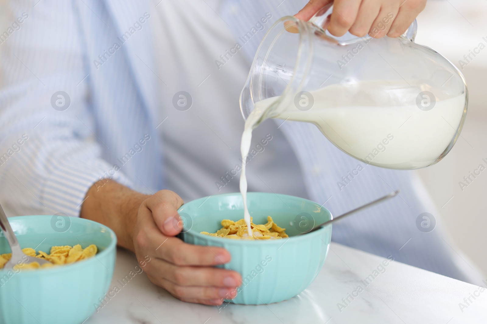 Photo of Making breakfast. Man pouring milk from jug into bowl with cornflakes at white marble table, closeup