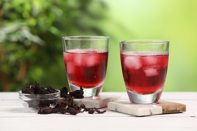 Refreshing hibiscus tea with ice cubes in glasses and dry roselle flowers on white wooden table against blurred green background