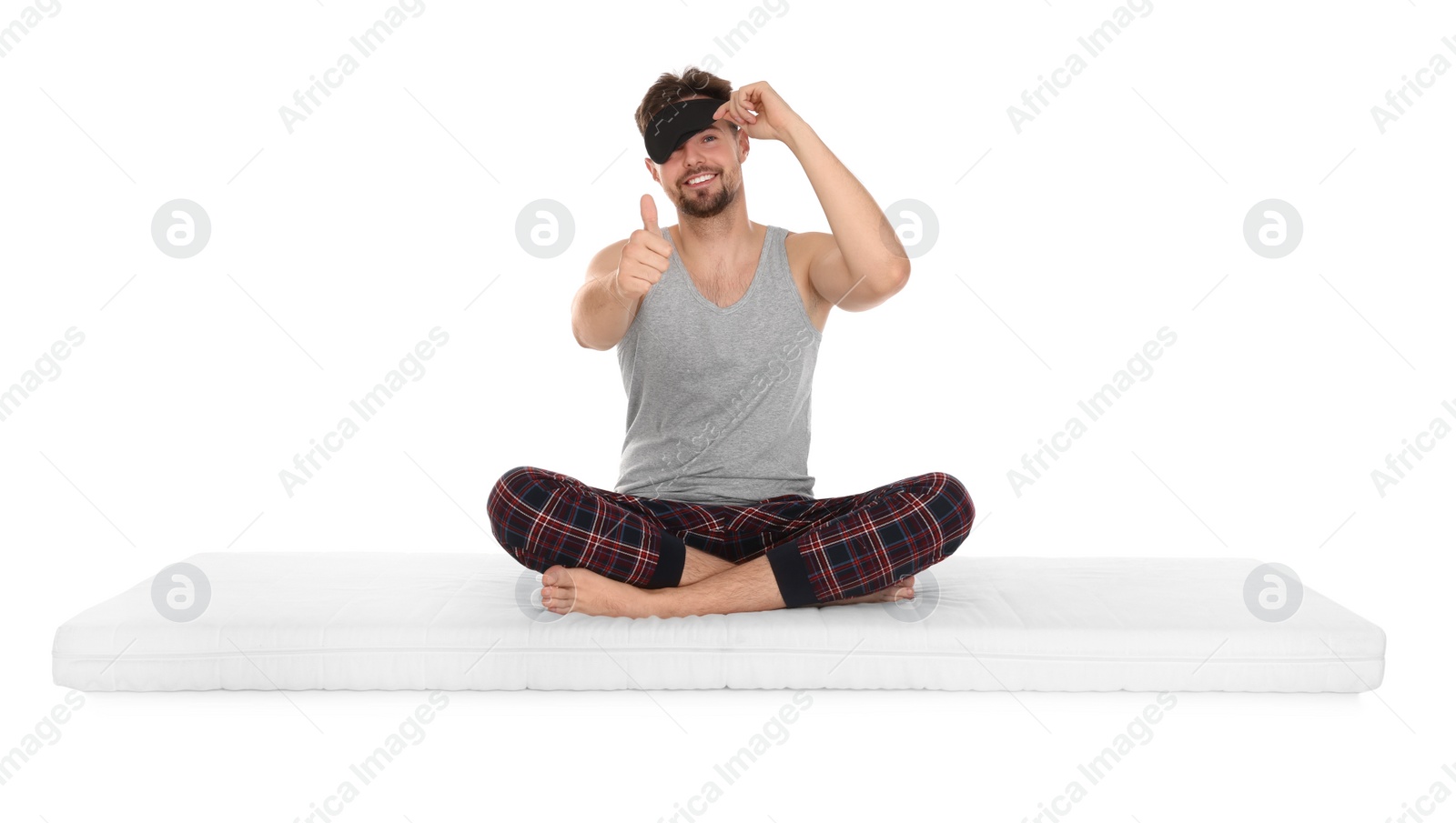 Photo of Smiling man in sleeping mask sitting on soft mattress and showing thumb up against white background