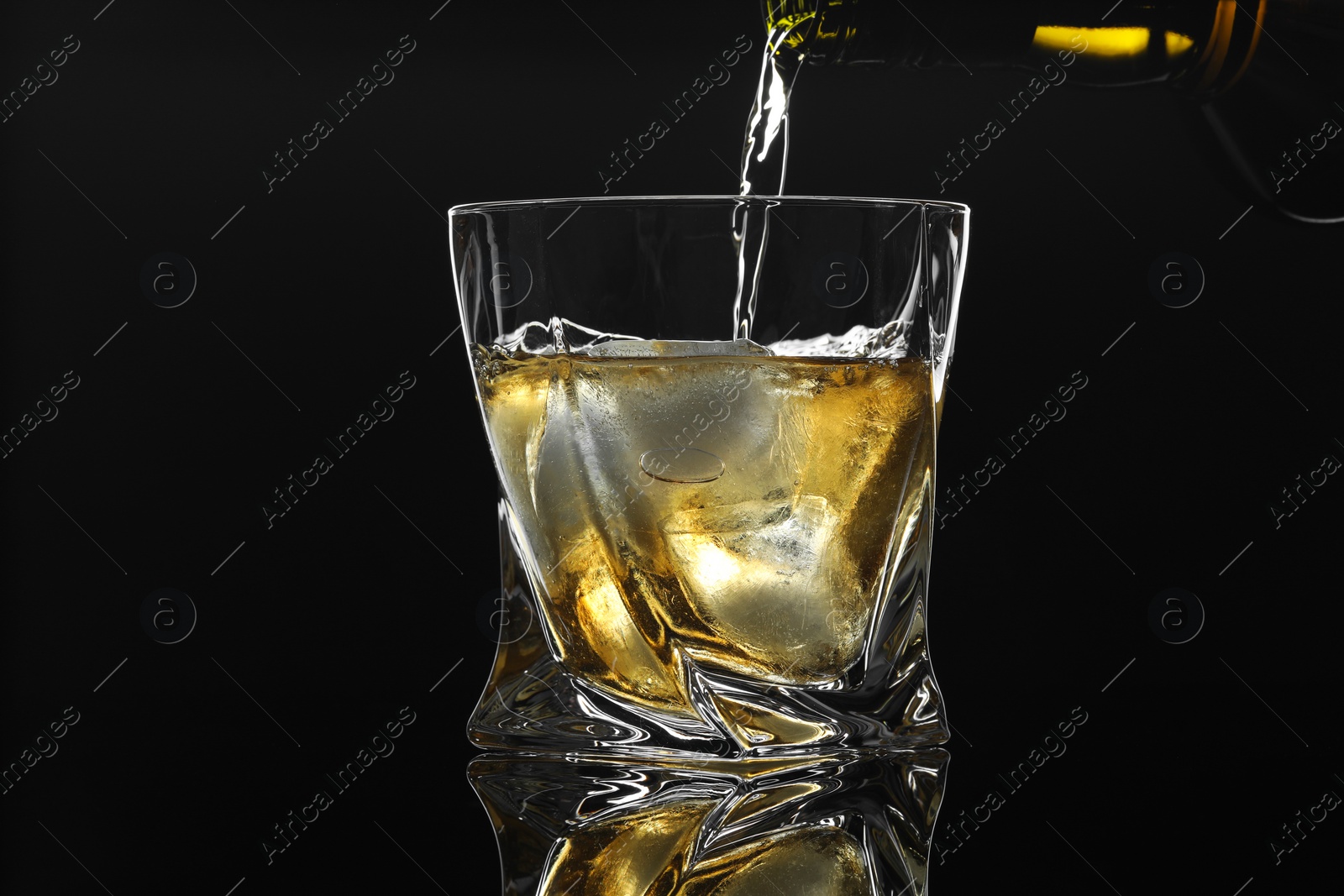 Photo of Pouring tasty whiskey from bottle into glass with ice at mirror table against black background, closeup