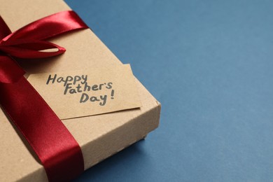 Photo of Card with phrase Happy Father's Day and gift box on blue background, closeup. Space for text