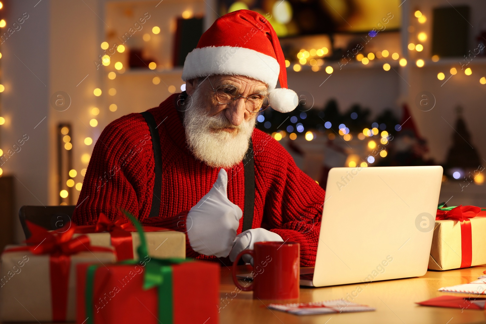 Photo of Santa Claus showing thumbs up near laptop at his workplace in room decorated for Christmas