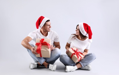 Photo of Beautiful happy couple in Santa hats sitting with Christmas gifts on light background