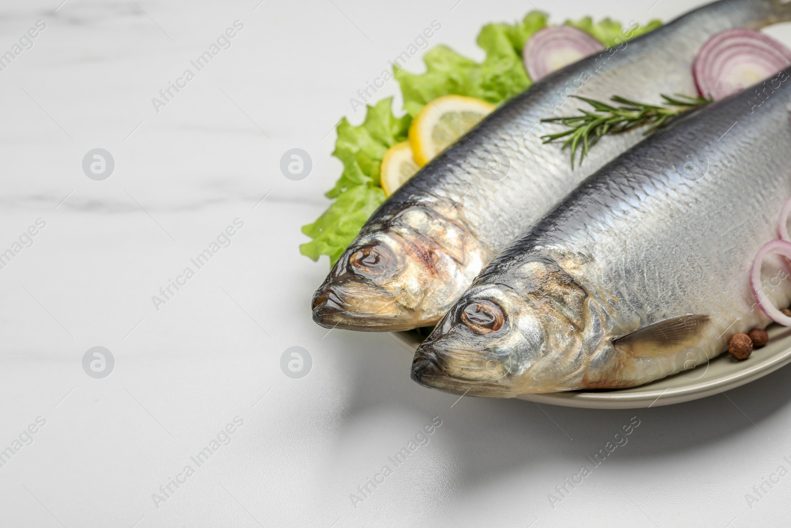 Photo of Plate with salted herrings, onion rings, slices of lemon, peppercorns and lettuce on white marble table, closeup. Space for text