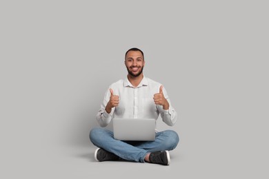 Smiling young man with laptop showing thumbs up on grey background