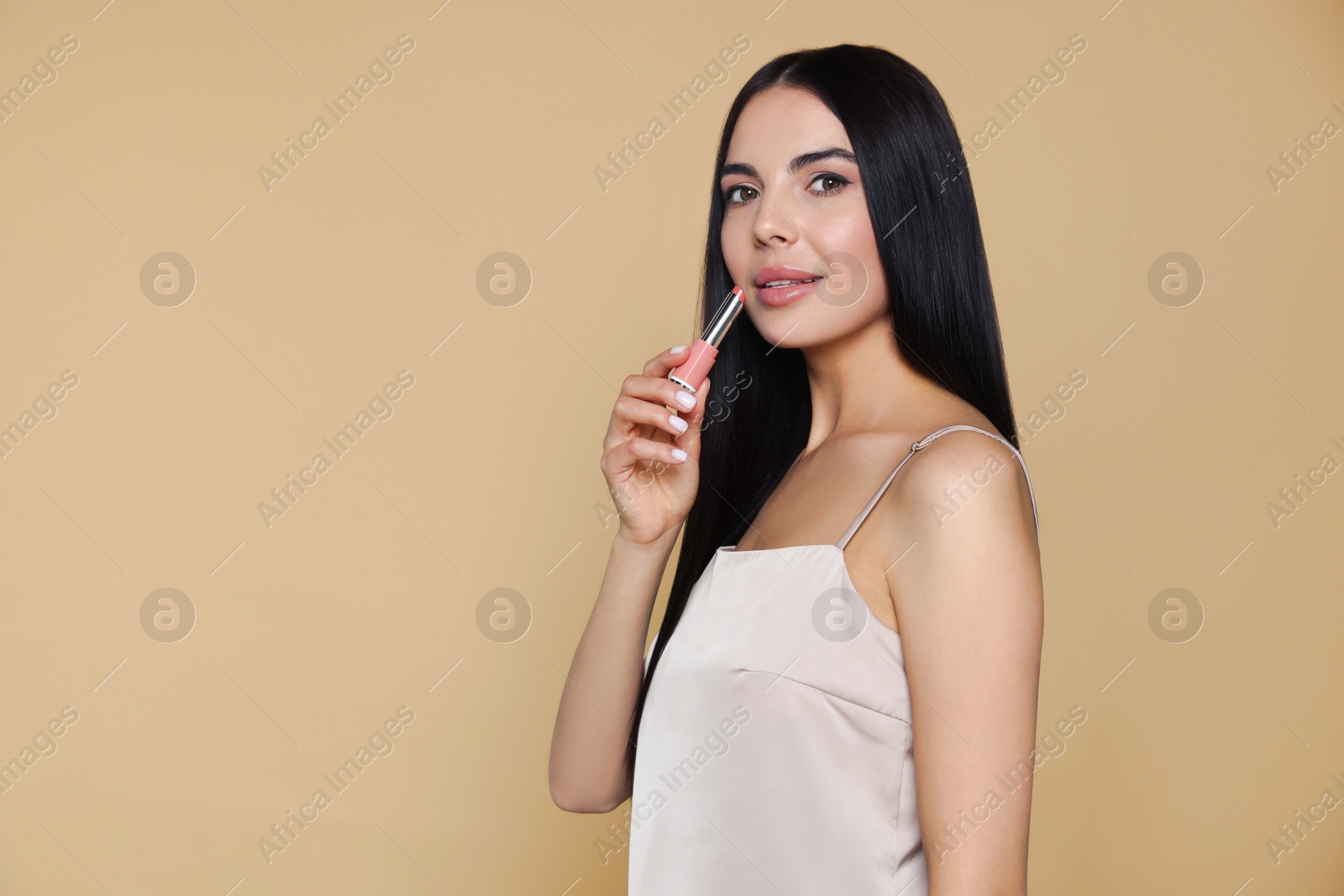 Photo of Young woman with beautiful makeup holding nude lipstick on beige background, space for text