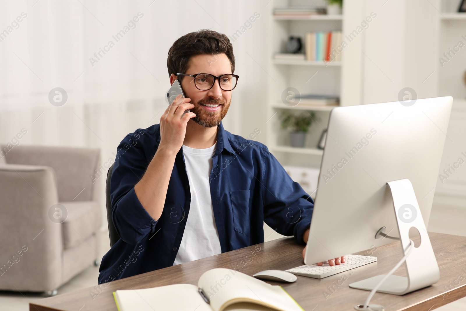 Photo of Home workplace. Happy man talking on smartphone while working with computer at wooden desk in room