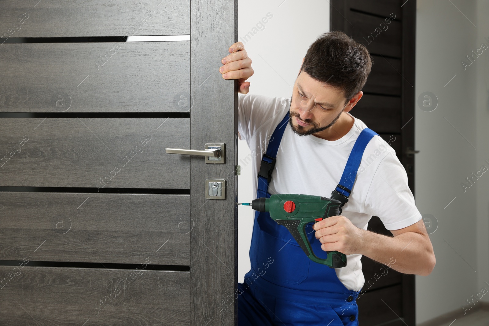 Photo of Worker in uniform with screw gun repairing door lock indoors