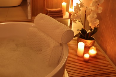 Photo of Tub with foamy water and soft bath pillow surrounded by candles indoors