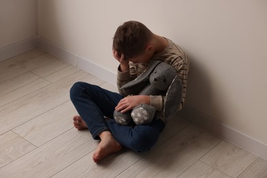 Photo of Child abuse. Upset boy with toy bunny sitting on floor near wall indoors